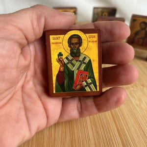 Op de afbeelding: Een klein, rechthoekig icoon dat Sint-Patrick van Ierland afbeeldt. Het icoon heeft een gouden halo, een groene en witte jurk en een rood boek. De heilige houdt een klaverblad vast. De tekst "SAINT CHRISTOPHER" en "OF IRELAND" is zichtbaar.