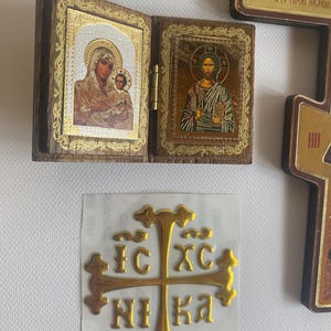 May include: A folding wooden diptych icon featuring a gold-toned image of the Virgin Mary and Child on the left and Jesus Christ on the right. Below, a gold-coloured cross sticker displays the Cyrillic text IC XC NI KA on a white backing.