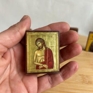 May include: A small, square religious icon depicting a figure in a red robe and gold halo. The icon is held in a hand and has a gold background. The figure has a crown of thorns and is holding a staff.