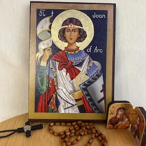 Puede incluir: Un icono de madera de Santa Juana de Arco, con fondo azul y detalles dorados. El icono representa a Juana de Arco con armadura, sosteniendo una espada y una bandera. El texto "St Joan of Arc" está escrito en el icono.
