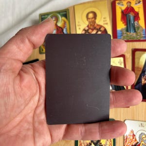 May include: A rectangular black magnet is held in front of a collection of religious icon cards. The cards feature images of saints and religious figures, with gold and colorful accents. The cards are arranged on a wooden surface.