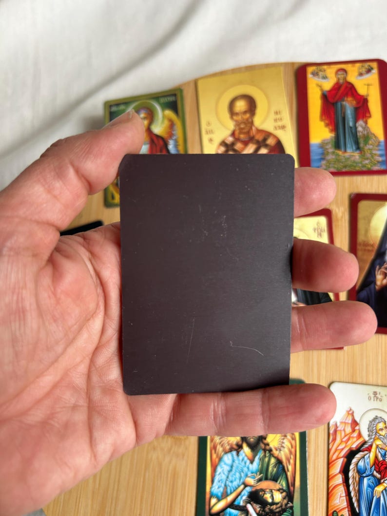 May include: A rectangular black magnet is held in front of a collection of religious icon cards. The cards feature images of saints and religious figures, with gold and colorful accents. The cards are arranged on a wooden surface.