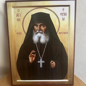 May include: A framed religious icon depicting a man with a long white beard and a black hooded robe. He wears a silver cross pendant and holds a rosary. The background is gold with Greek text and the word "ARIZONA".