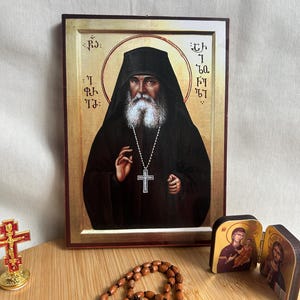 May include: An icon depicting a religious figure with a long white beard, wearing a black robe and a cross necklace. A wooden rosary, a small cross, and a miniature icon are also present.