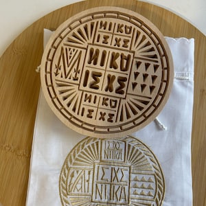 May include: A wooden, circular carving and a gold embroidered patch with Greek text. The wooden carving is light brown with a geometric design and the Greek text "NIKA". The embroidered patch is gold on a white background.