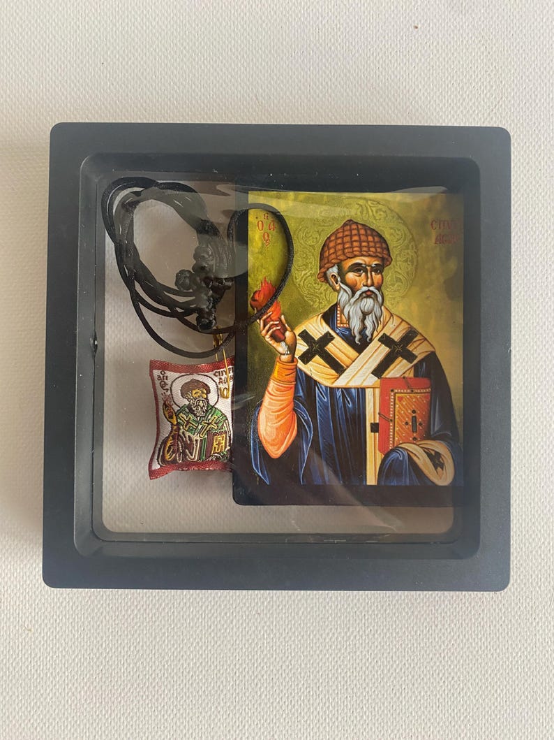 May include: A framed religious icon featuring a bearded figure in a blue robe, holding a book and a flame. A small pendant with a similar image and a black cord are also included. The frame is black.