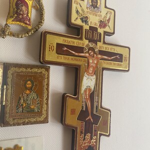 May include: A wooden Orthodox cross featuring a central depiction of the crucifixion with Cyrillic text, accompanied by a small hinged wooden icon of Christ and a gold-toned woven bracelet with a small square icon of the Virgin Mary and Child.