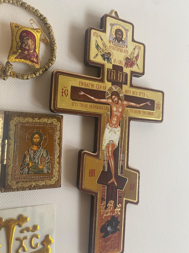 May include: A wooden Orthodox cross featuring a central depiction of the crucifixion with Cyrillic text, accompanied by a small hinged wooden icon of Christ and a gold-toned woven bracelet with a small square icon of the Virgin Mary and Child.