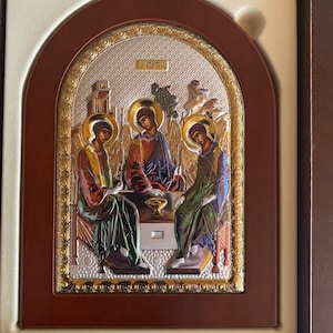 Puede incluir: Icono religioso que representa tres figuras con halos, sentadas alrededor de una mesa. La obra de arte presenta detalles dorados y plateados, enmarcada en madera. Las figuras visten túnicas de varios colores, con elementos arquitectónicos de fondo.