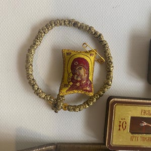 May include: A gold-coloured knotted cord bracelet featuring a small, rectangular, embroidered fabric icon of the Virgin Mary and Child. The icon is yellow with red details and includes the letters MP and OY. A gold safety pin is attached to the top.