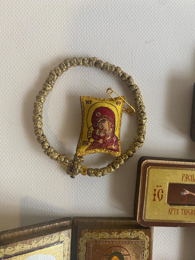 May include: A gold-coloured knotted cord bracelet featuring a small, rectangular, embroidered fabric icon of the Virgin Mary and Child. The icon is yellow with red details and includes the letters MP and OY. A gold safety pin is attached to the top.
