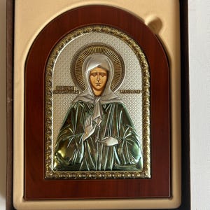 May include: Religious icon featuring a woman in a green robe and white head covering, set against a silver and gold background. The icon is framed in dark wood and presented in a beige box.