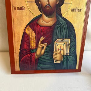 Może przedstawiać: Ikona religijna przedstawiająca Jezusa Chrystusa trzymającego książkę. Ikona jest namalowana w tradycyjnym stylu z żywymi kolorami, w tym czerwonym, zielonym i złotym. Tło jest jasnoż&oacute;łte z złotymi akcentami. Tekst "O ПАНТО КРАТОР" jest napisany po grecku u g&oacute;ry ikony.