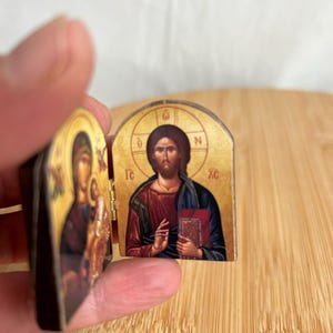 May include: A small, hinged diptych icon featuring religious imagery. The right panel depicts a figure in a red robe and blue cloak holding a book, with the Greek letters IC XC visible. The left panel shows a figure holding a child.