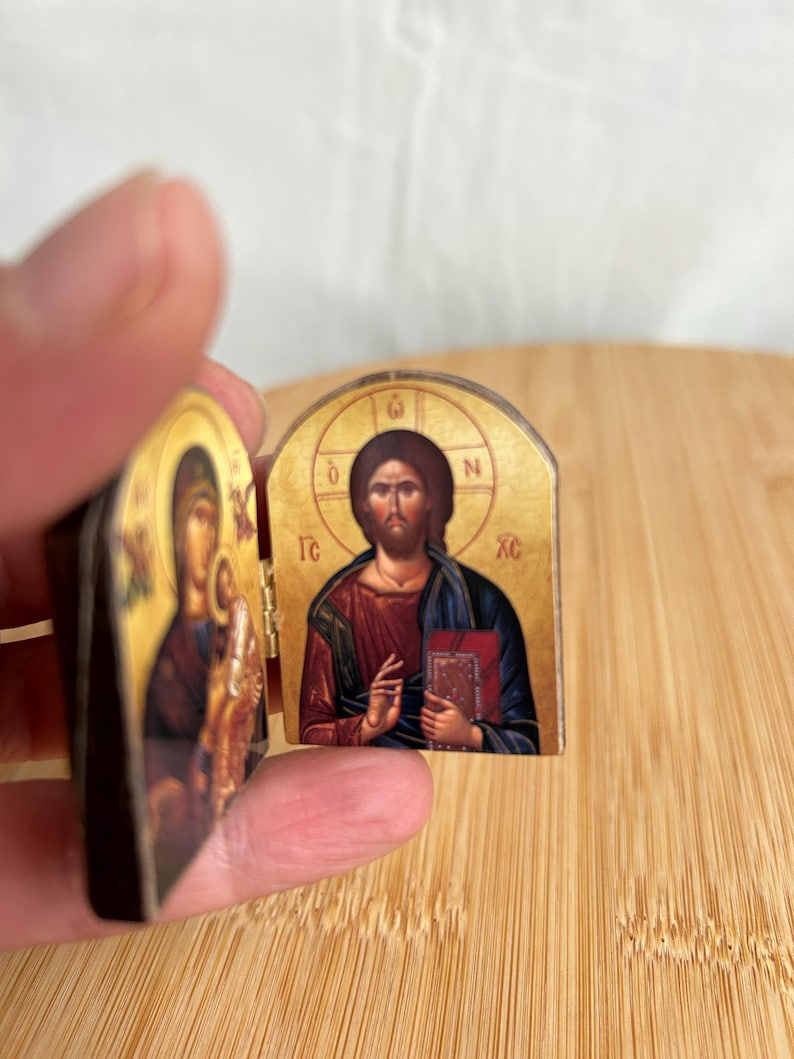 May include: A small, hinged diptych icon featuring religious imagery. The right panel depicts a figure in a red robe and blue cloak holding a book, with the Greek letters IC XC visible. The left panel shows a figure holding a child.