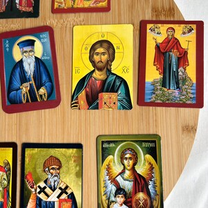 May include: A collection of religious icon cards featuring various saints and biblical figures. The cards have a rectangular shape with a red border. The images depict detailed illustrations with vibrant colors, including gold, blue, and red. The cards are arranged on a wooden surface.