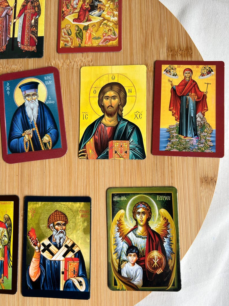 May include: A collection of religious icon cards featuring various saints and biblical figures. The cards have a rectangular shape with a red border. The images depict detailed illustrations with vibrant colors, including gold, blue, and red. The cards are arranged on a wooden surface.
