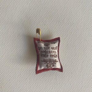May include: Small, rectangular religious icon with gold trim and text in a foreign language, attached to a gold safety pin. The icon is white with red trim and a decorative border. The text is in gold.