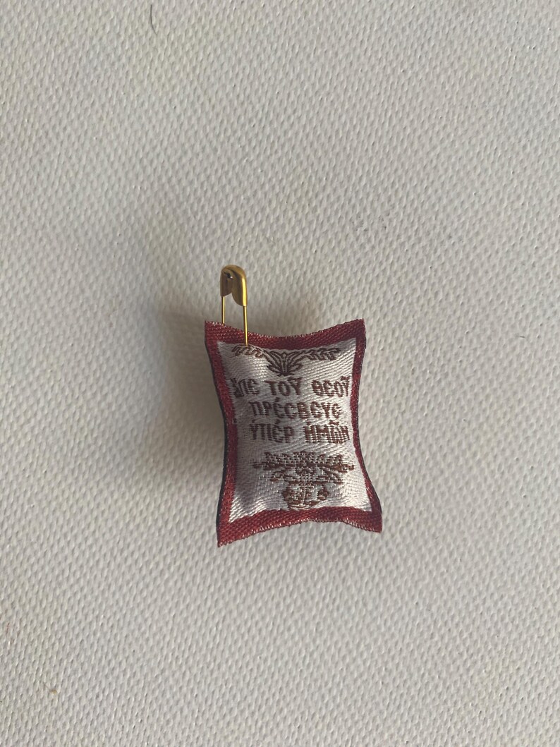 May include: Small, rectangular religious icon with gold trim and text in a foreign language, attached to a gold safety pin. The icon is white with red trim and a decorative border. The text is in gold.