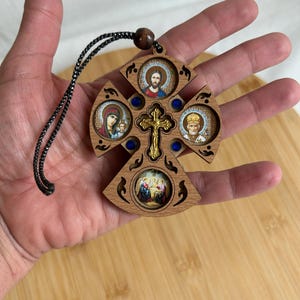 May include: Wooden cross pendant featuring four religious icons and a gold-coloured central cross. The cross has blue accents and a black cord. The pendant is held in a hand.