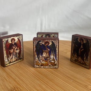 May include: Several small, rectangular wooden blocks with colorful illustrations of angels. The blocks feature images of St. Michael and St. Raphael, with detailed artwork and text. The blocks are displayed on a wooden surface.
