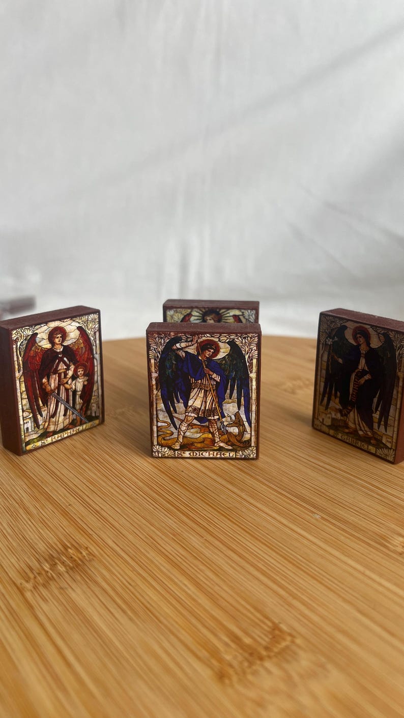 May include: Several small, rectangular wooden blocks with colorful illustrations of angels. The blocks feature images of St. Michael and St. Raphael, with detailed artwork and text. The blocks are displayed on a wooden surface.