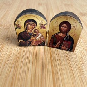 May include: A small, hinged diptych icon featuring two arched panels. The left panel depicts a figure holding a child against a gold background, while the right panel shows a bearded figure in red and blue robes holding a book, also on gold.