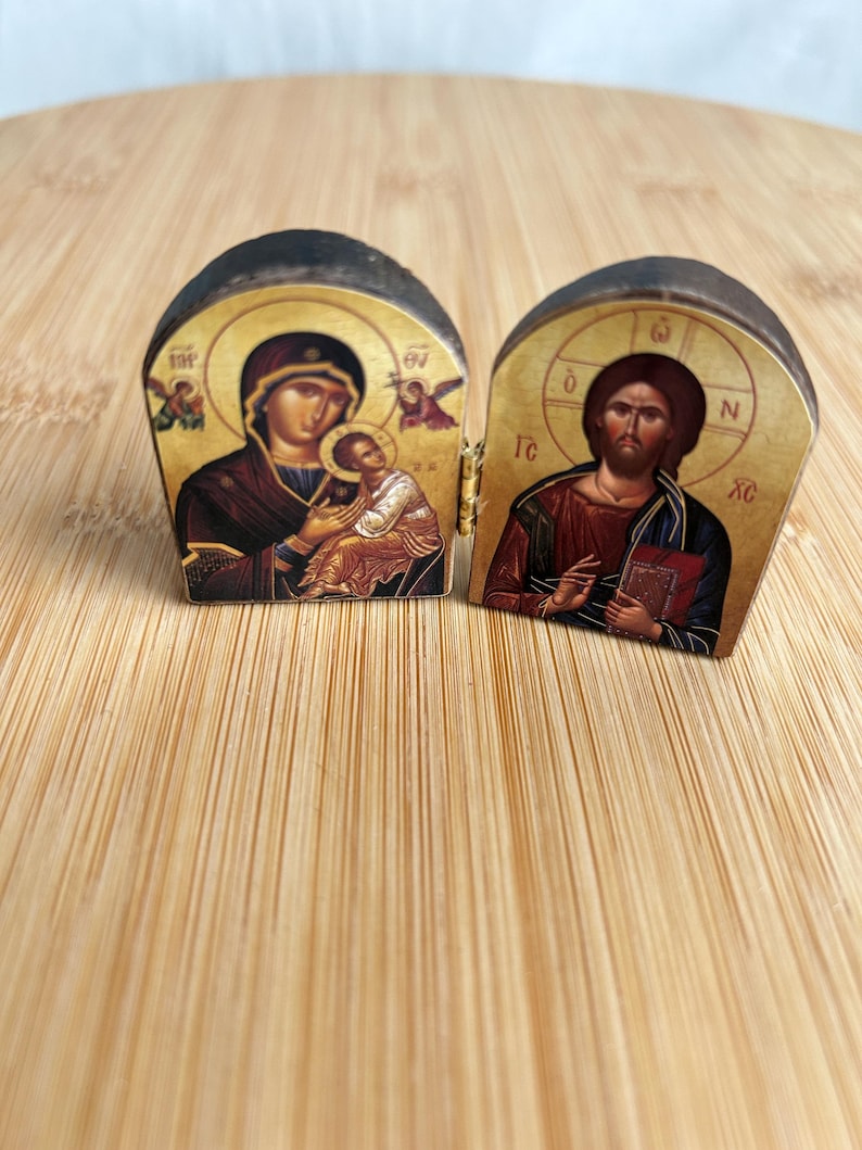 May include: A small, hinged diptych icon featuring two arched panels. The left panel depicts a figure holding a child against a gold background, while the right panel shows a bearded figure in red and blue robes holding a book, also on gold.
