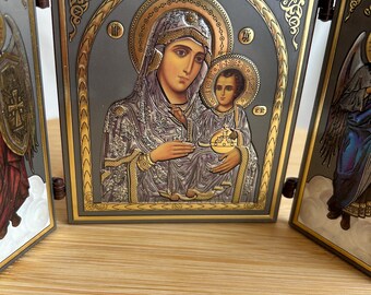 Könnte beinhalten: Ein Triptychon-Ikonensatz mit goldener und silberner Oberfläche. Das mittlere Feld zeigt die Jungfrau Maria, die das Jesuskind hält. Die linken und rechten Felder zeigen Engel mit Flügeln.