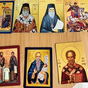 May include: A collection of religious icon cards featuring various saints and religious figures. The cards have gold backgrounds and depict figures in traditional attire, with some holding books or scrolls. The images are colorful and detailed.