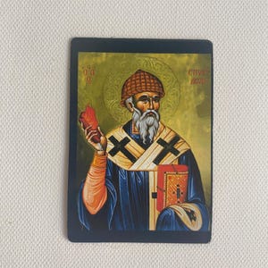 May include: Rectangular religious icon featuring a saint with a long white beard, wearing a blue robe and a brown hat. The saint holds a red object in one hand and a book in the other. The background is gold with text.