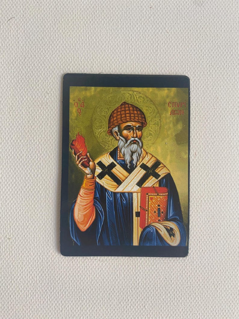 May include: Rectangular religious icon featuring a saint with a long white beard, wearing a blue robe and a brown hat. The saint holds a red object in one hand and a book in the other. The background is gold with text.