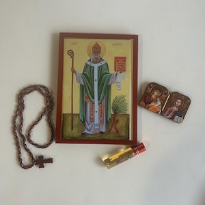 May include: A wooden framed icon of Saint Martin with a gold background and a green robe. The icon is accompanied by a brown wooden rosary with a cross, a small wooden icon of the Holy Family, and a small bottle of oil.