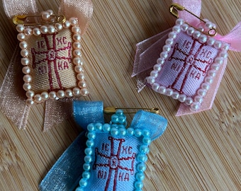 May include: Three small embroidered squares with a cross and text in red thread, surrounded by white pearls and tied with ribbons in pink, blue and peach.