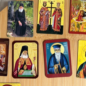 May include: A collection of religious icons featuring images of saints and religious figures. The icons are rectangular with various colors and designs, including images of people, crosses, and religious symbols. They are arranged on a light brown wooden surface.