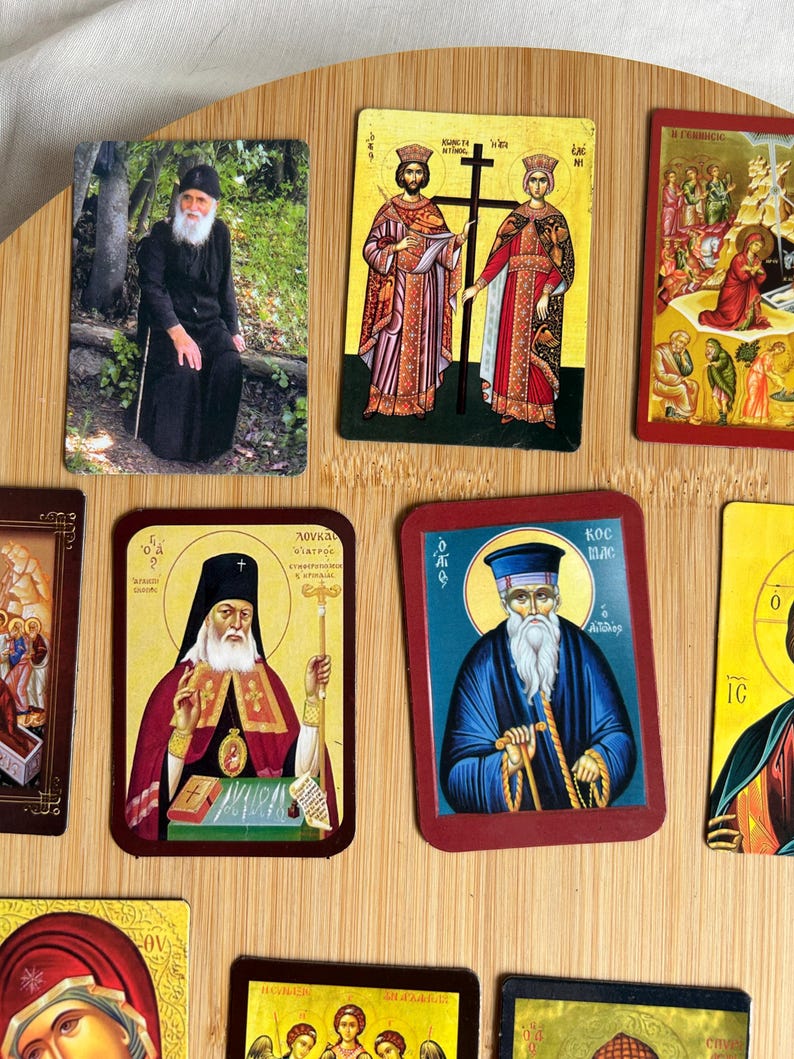 May include: A collection of religious icons featuring images of saints and religious figures. The icons are rectangular with various colors and designs, including images of people, crosses, and religious symbols. They are arranged on a light brown wooden surface.
