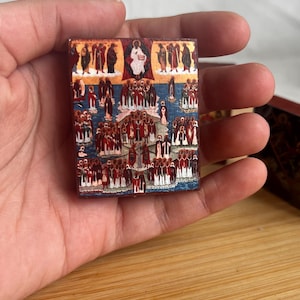 May include: A small, rectangular religious icon featuring a detailed depiction of a biblical scene. The artwork showcases figures in red, white, and blue robes, set against a blue background. The icon is held in a hand, suggesting its small size and portability.