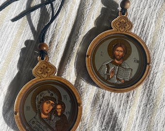 May include: Two wooden religious pendants with black cords. One pendant features an image of Jesus holding a book, the other depicts Mary and the baby Jesus. The pendants have a light brown wooden frame and a decorative top.
