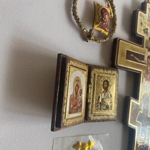 May include: A gold-toned beaded bracelet with a small religious charm, a wooden diptych icon featuring depictions of the Virgin Mary and Jesus, a large wooden crucifix, and a gold-coloured sticker with the letters IC XC on a white surface.