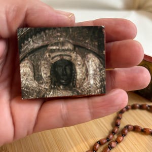 May include: A small, rectangular religious icon depicting a crowned figure with wings. The icon is held in a hand, with a wooden bead necklace in the foreground. The icon has a vintage appearance. The image is on a wooden surface.