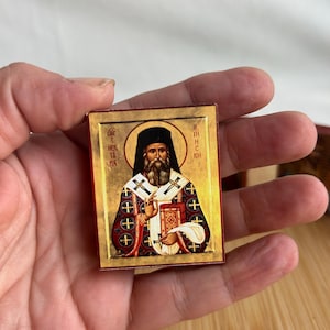 May include: A small, rectangular icon depicting a religious figure with a long beard, wearing a black hat and ornate robes. The icon has a gold background and includes text in a foreign language. The icon is held in a hand.