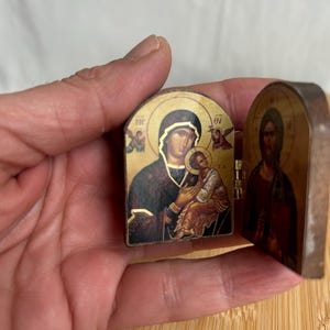 May include: A small, hinged, diptych-style religious icon featuring a gold-toned background. The left panel depicts a figure holding a child, while the right panel shows a bearded figure. Both panels include small, dark-coloured text characters.