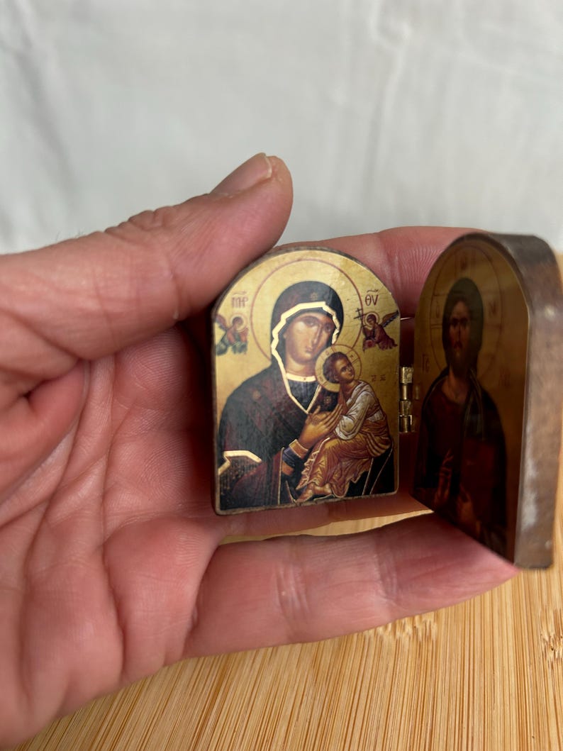 May include: A small, hinged, diptych-style religious icon featuring a gold-toned background. The left panel depicts a figure holding a child, while the right panel shows a bearded figure. Both panels include small, dark-coloured text characters.
