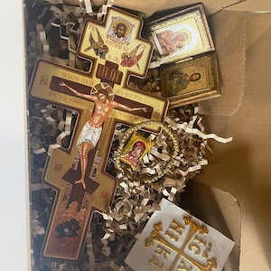 May include: A cardboard box containing a large wooden crucifix with a depiction of the crucifixion, two small rectangular religious icons, a gold-toned beaded bracelet with a small religious pendant, and a square card featuring a gold cross design.