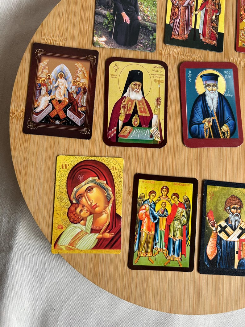 May include: A collection of religious icon cards featuring various saints and biblical scenes. The cards display vibrant colors and intricate details, with gold accents and ornate borders. The images are set against a light wooden background.