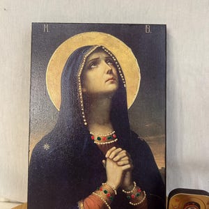 May include: A religious icon depicting the Virgin Mary with a sorrowful expression, wearing a dark blue robe with gold and green accents. The icon is framed in a dark wood frame.