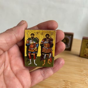 Pode incluir: Um pequeno ícone de madeira com fundo dourado que representa duas figuras com armadura segurando lanças. O ícone tem uma borda vermelha e o texto "ΑΓΙΟΙ ΑΝΑΡΓΥΡΟΙ" em grego.