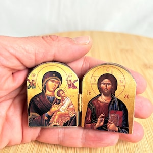 May include: A small, hinged diptych icon featuring religious imagery on two wooden panels. The left panel depicts a figure holding a child, while the right panel shows a bearded figure holding a book. Both panels feature gold-toned backgrounds.