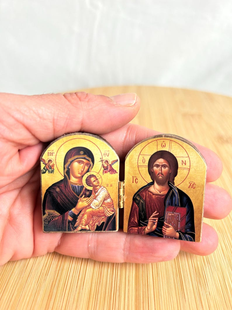 May include: A small, hinged diptych icon featuring religious imagery on two wooden panels. The left panel depicts a figure holding a child, while the right panel shows a bearded figure holding a book. Both panels feature gold-toned backgrounds.