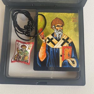 May include: A religious icon and pendant set in a black display case. The icon depicts a saint holding a flame and a book, with text in a foreign language. A small pendant with a similar image hangs from a black cord.